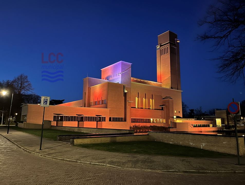 The town hall at night against a dark sky, bathed in shades of red, orange and purple, the shadows created by the illumination serving to emphasise the linear nature of the town hall’s different components (for example, there is a strong contrast between the brightly-lace of one side of the campanile and the shaded neighbouring face).