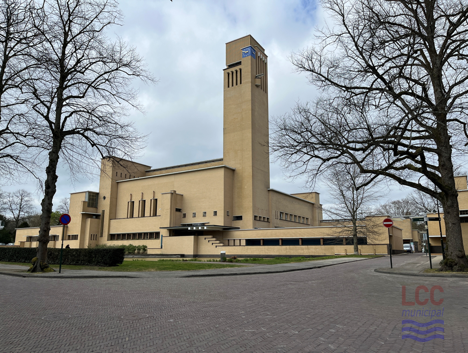 An exterior shot of Hilversum Town Hall taken in daylight. The building is composed of various asymmetrical cuboid blocks in yellow brick, dominated by a tall chimney-like clock tower