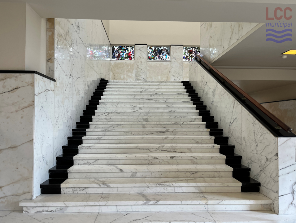 The wide staircase is of a white marbled material with black edges at either side. At the top is a landing before the staircase continues its ascent in the opposite direction. Some rectangular stained glass windows can be seen at the top of the stairs.