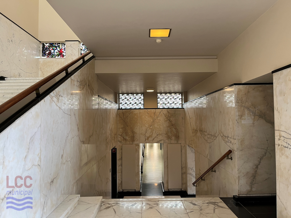 The floor and walls are of a white marbled material. The view looks down the stairs to a long corridor at their foot. The the left can be seen the main ceremonial staircase rising to the Council Chamber and Assembly Hall.
