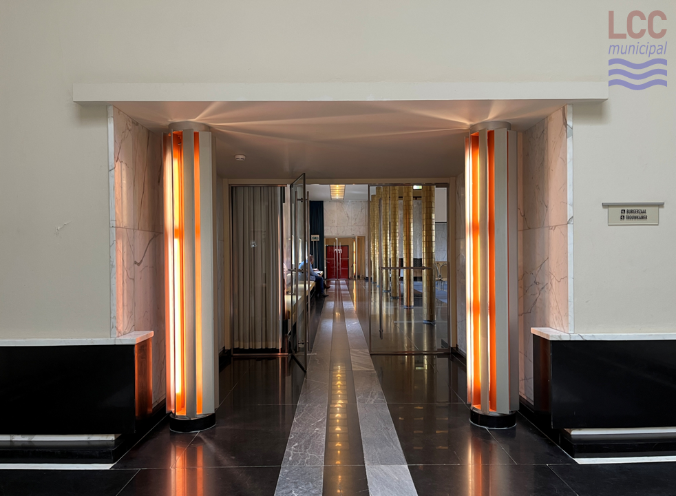 Either side of the entrance are two illuminated columns, giving off an orange light. The floor is of black and grey tiles which reflect the ceiling lights. Two glass doors invite people into the hall.