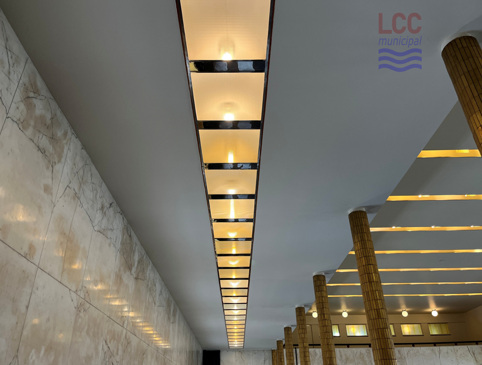 A long rectangular ceiling lighting unit recedes into the distance. On the left hand side is a marbled wall. On the right hand side a row of columns can be seen, covered in gold-coloured tiles.