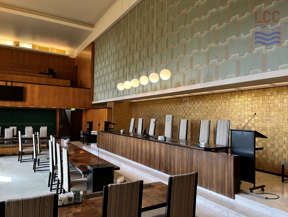 Six globe lights hang from the ceiling. Beneath them are the tables at which the Councillors sit. The chairs are high-backed and covered in a grey fabric. They face a raised area, where the Council cabinet sits behind a desk, facing the Councillors. There is a chequerboard-effect carpet in cream and green. In the distance, a raised public gallery can be seen.