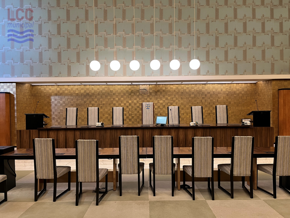 Six globe lights hang from the ceiling. Beneath them are the tables at which the Councillors sit. The chairs are high-backed and covered in a grey fabric. They face a raised area, where the Council cabinet sits behind a desk, facing the Councillors. There is a chequerboard-effect carpet in cream and green.