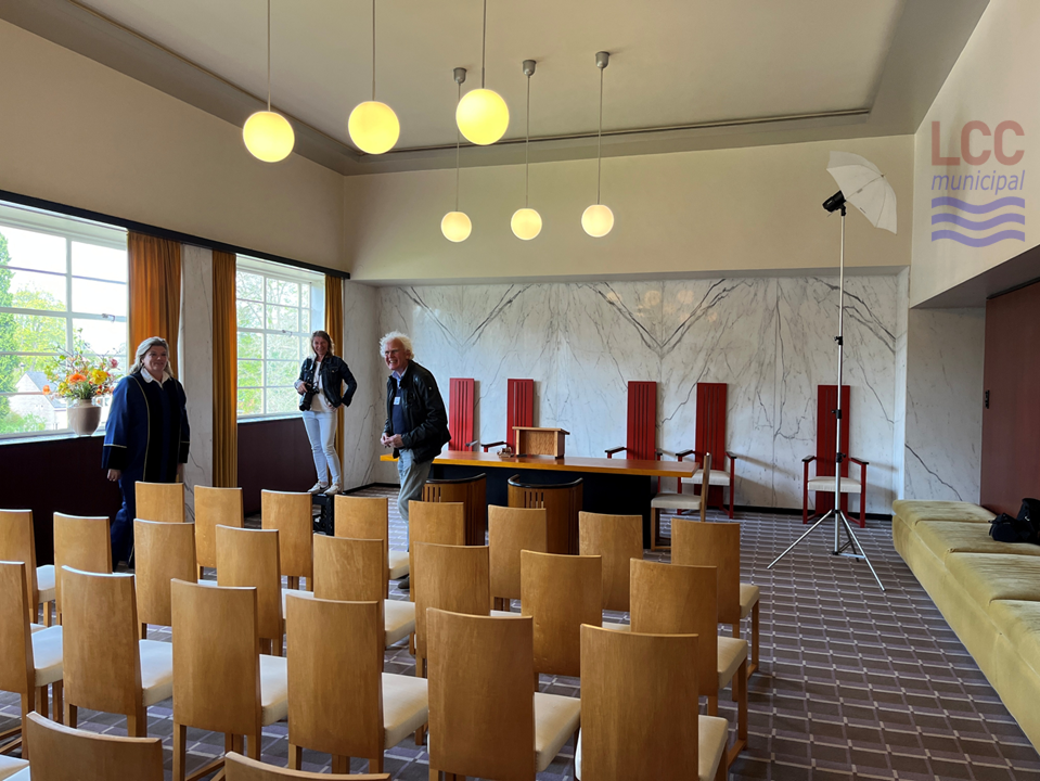 A large room with a grey/purple carpet with a geometric pattern. High-backed wooden chairs are arranged in rows on the carpet, facing towards the ceremonial area at the front of the room. At the front of the room, high backed chairs in red are behind a large wooden desk, facing the audience. Globe-shaped lights hang from the ceiling.