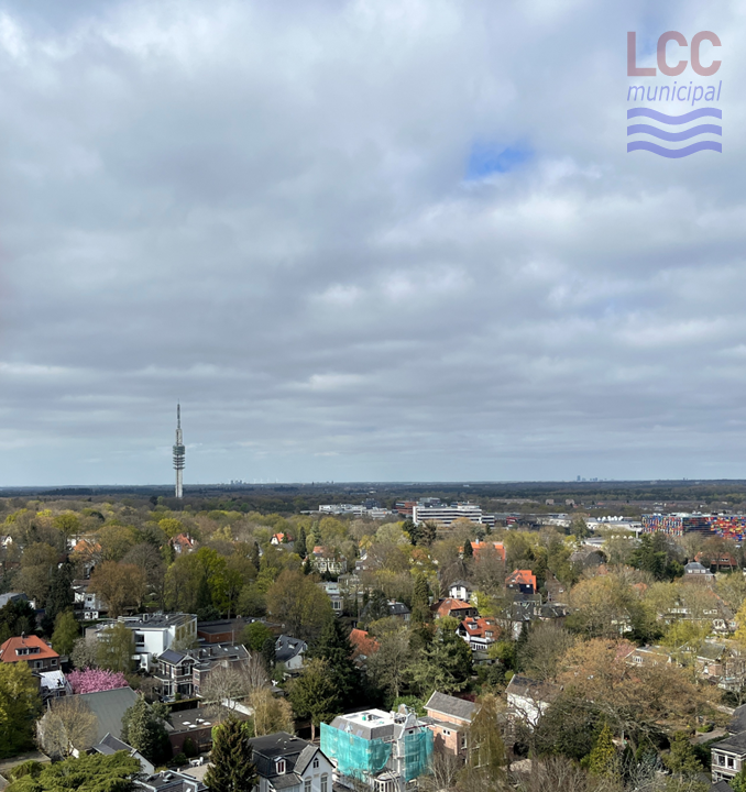 A view looking north from the top of the bell tower. In the foreground are suburban houses set among trees and greenery. In the middle distance is Hilversum's media park with a tall transmitter dominating the scene.