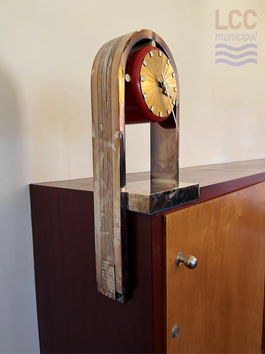 A clock attached to the side of a cabinet. The round clock face sits in the bend of a U-shaped metal frame. The edge of the clock is red and the face is golden in colour.