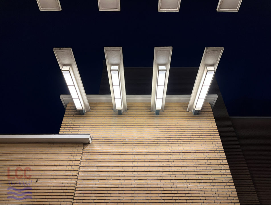 Four bars stick out of the side of a yellow brick wall, like the prongs of a rake. On each is a long cuboid lighting unit in a slight Art Deco style. As it is nighttime, they are illuminated and give off a bright white light which contrasts with the dark sky behind.