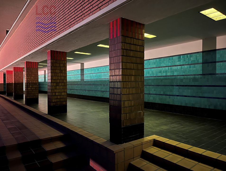 One of the walkways up to the front of the building. It runs alongside a large rectangular pond. It is a covered walkway with rectangular inset lighting blocks receding into the distance. Bands of blue tiles line one side of the walkway.