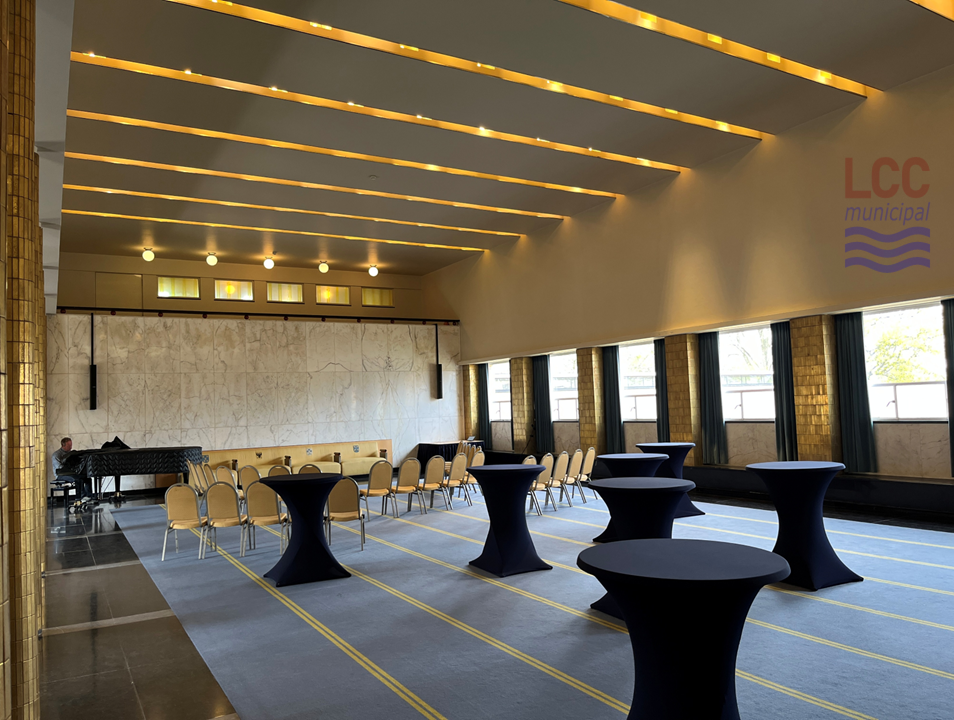 A large hall with a blue carpet with a thin yellow stripe. Tables and chairs are scattered around the place. There is a bank of windows on the right hand side and just visible on the left hand side is a row of columns with gold tiles. The ceiling has banks of lights arranges in long rows running across the ceiling. The striped effect of the lighting echoes the stripe on the carpet but is at 90 degrees to it, so they run in different directions.