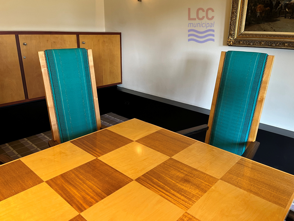 Two chairs behind a table. The chairs have high backs and are covered in a blue fabric. The table is wooden with a chequerboard pattern of light and dark wood.