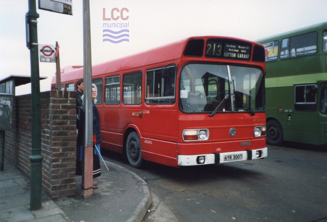 LS300 is a single deck Leyland National red London Transport bus. It stands in the old bus yard at Kingston Station. A green London Country Leyland Atlantean double decker can be seen in the background. People queue to board the bus.