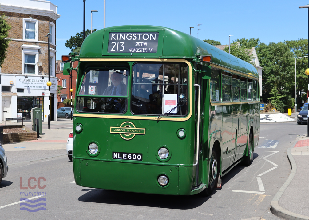 RF600 is a green London Transport Country area single deck bus from the 1950s. It has a distinctive rounded and stream-lined shape, making it look very modern at the time. It is pictured in an urban scene with a roundabout, trees and shops in the background.