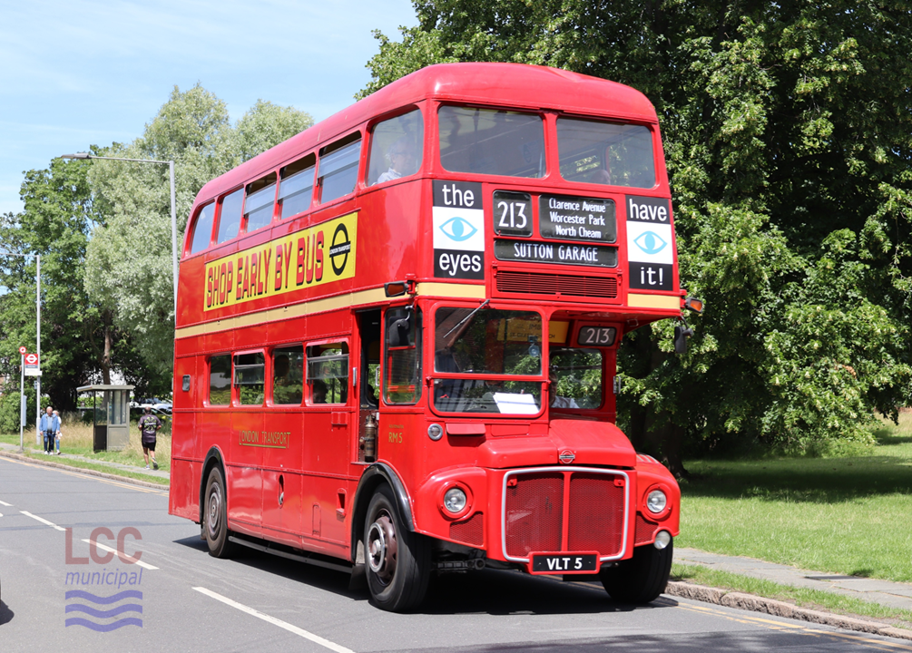 RM 5, a red double deck front-engine rear-entrance bus. It is travelling toward the camera on a suburban street with large trees behind. The side of the bus carries a large advertisement saying "Shop Early by Bus"