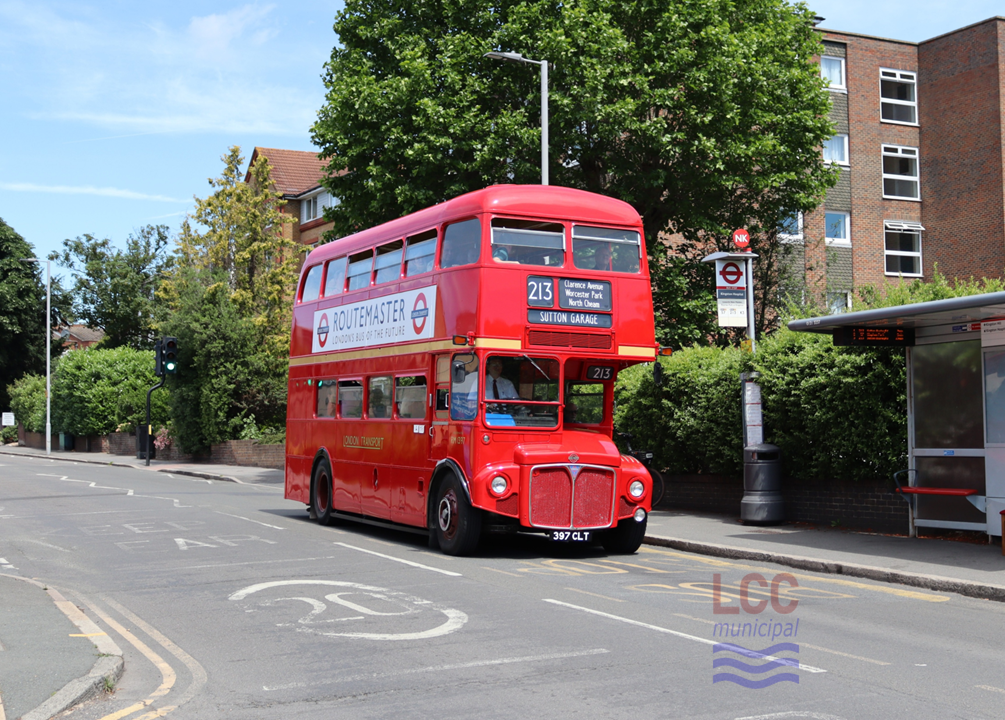 RM1397, a red double deck front-engine rear-entrance bus. It stands by a bus stop on a suburban street with low rise blocks of flats and trees behind. The side of the bus carries a large advertisement saying "Routemaster London's Bus of the Future"