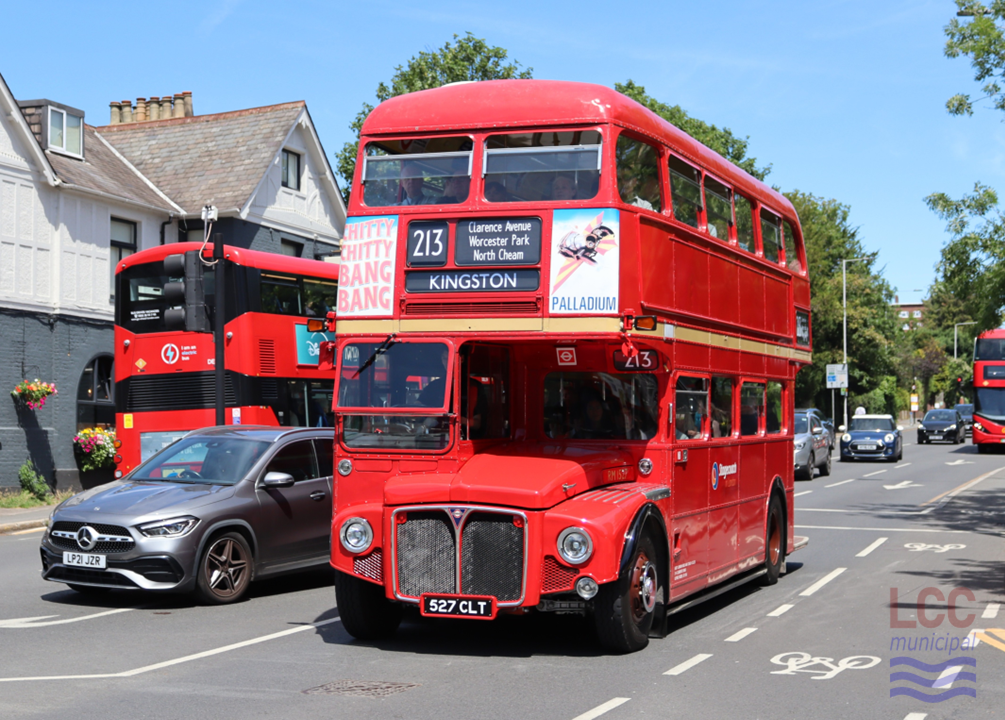 RM1527, a red double deck front-engine rear-entrance bus. It is travelling toward the camera on Kingston Hill with a pub, trees and other buses visible behind on this busy street. The front of the bus carries advertisements for the stage version of Chitty Chitty Bang Bang. It carries Stagecoach fleet names.