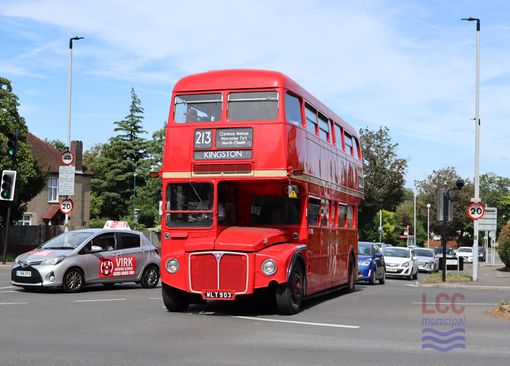 RM903, a red double deck front-engine rear-entrance bus. It is crossing the Sutton bypass at the head of a queue of cars, with houses and trees visible behind. The bus does not carry any advertising.